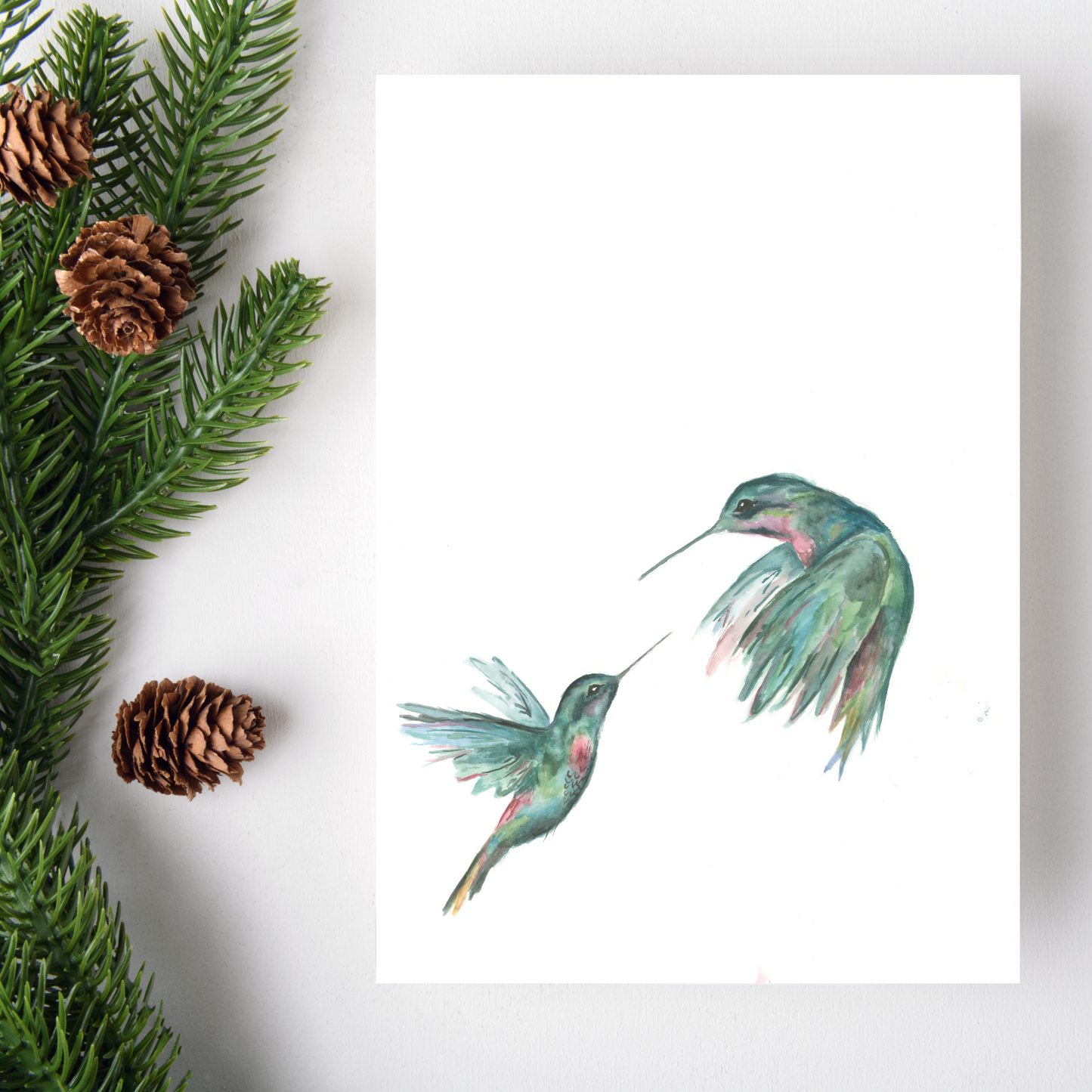 Two watercolor hummingbirds on a white background with greenery and pine cones.