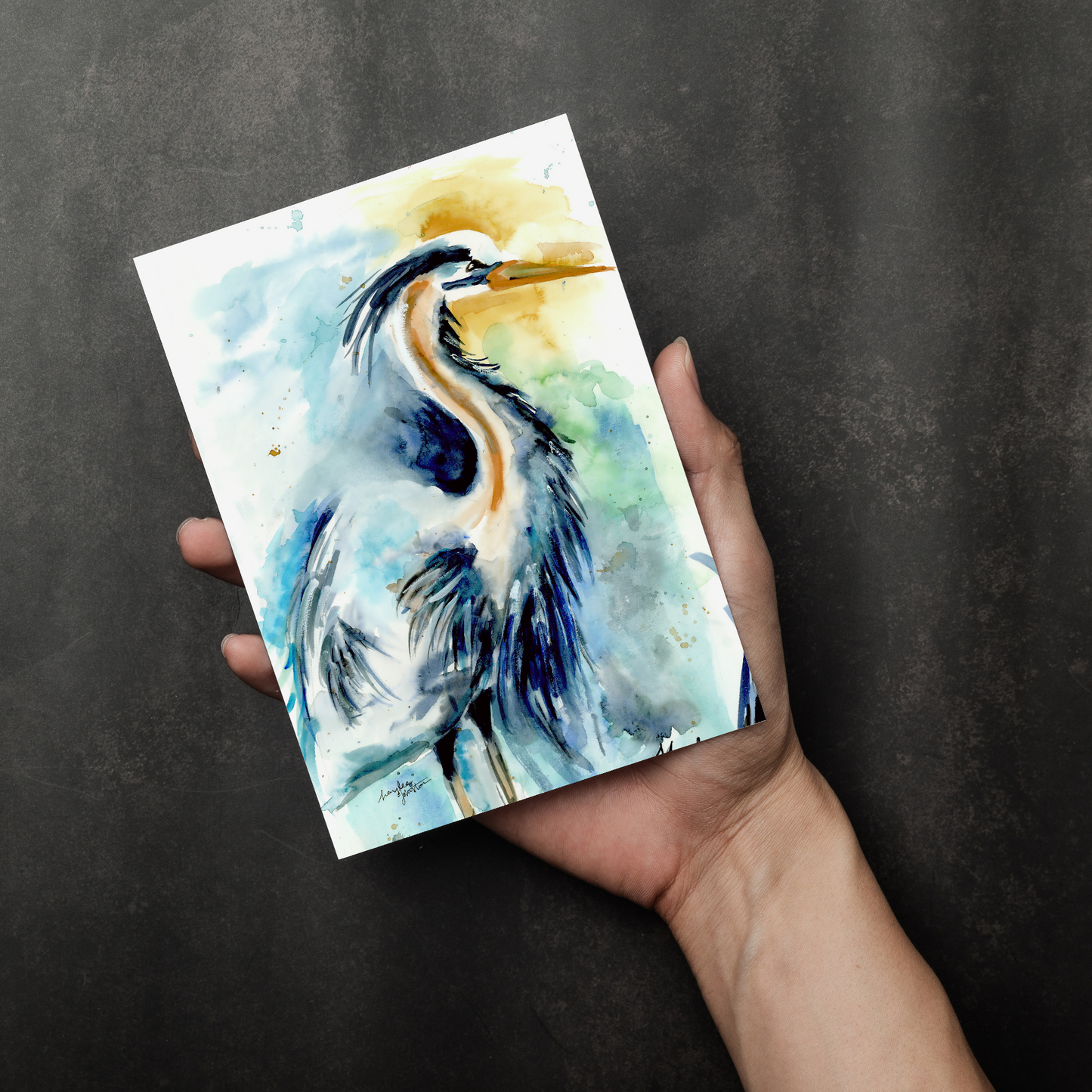 Flat cardstock note card featuring original watercolor piece Male Heron in a person's right hand. The background transitions from black at the bottom to a dull gray at the top.