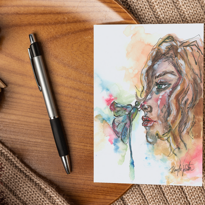 Flat cardstock note card featuring original watercolor piece Dragonfly Girl displayed on a wooden surface beside a black and silver pen.