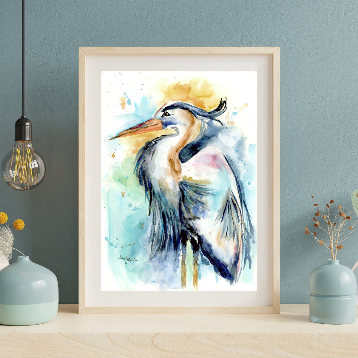 Giclée Female Heron in a light wood frame with white matting leans against a dusty blue textured wall on a pale wood surface. An exposed Edison bulb pendant hangs to the left, and soft blue ceramic vases with dried botanicals sit on either side.