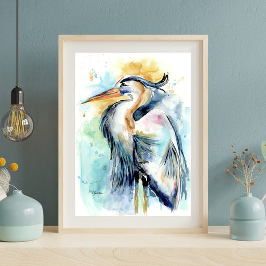 Giclée Female Heron in a light wood frame with white matting leans against a dusty blue textured wall on a pale wood surface. An exposed Edison bulb pendant hangs to the left, and soft blue ceramic vases with dried botanicals sit on either side.
