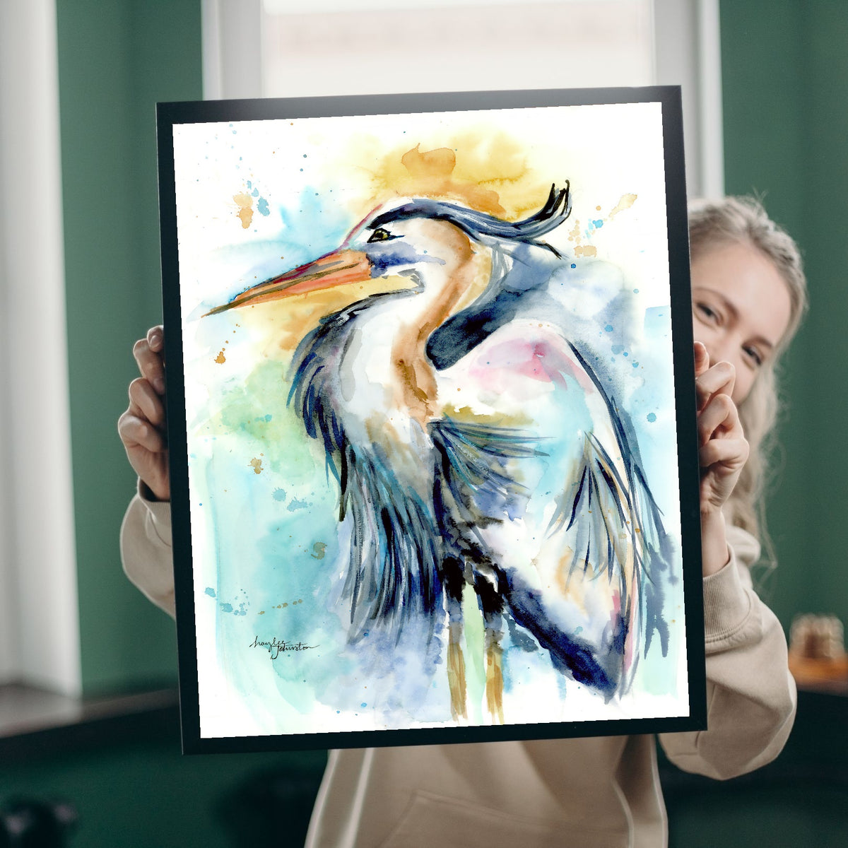 A smiling woman with blonde hair peeks out from behind Giclée Female Heron, holding it up in a black frame in a room with green walls and bright windows, giving a sense of the print's real-life scale.