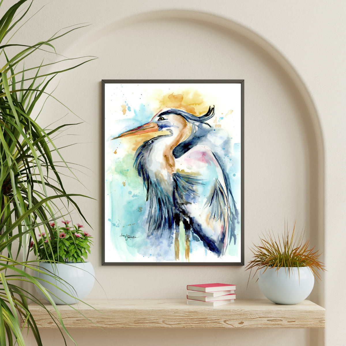 Giclée Female Heron in a thin black frame hangs inside an arched alcove on a cream wall, above a chunky light wood shelf styled with a stack of pink books and two round white pots holding green and bronze plants.