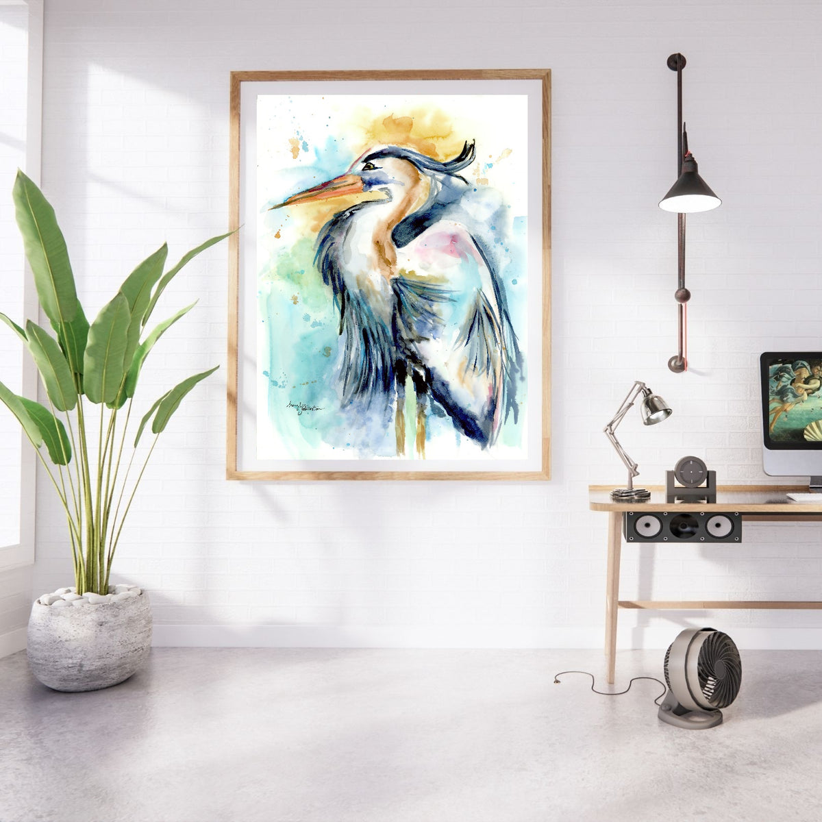 Giclée Female Heron in a large oak frame with white matting hangs on a white brick wall in a bright studio. A tall leafy plant in a stone planter stands to the left, and a light wood desk with a computer monitor, adjustable lamp, and speaker sits to the right beneath an industrial wall light.