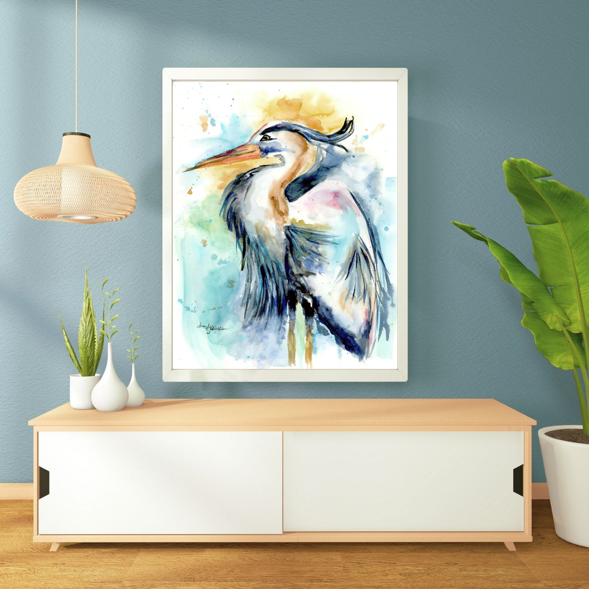 Giclée Female Heron in a white frame hangs on a soft blue-teal wall above a low light wood console with white sliding doors. A woven rattan pendant lamp hangs to the left, small white vases with greenery sit on the console, and a tall banana leaf plant stands to the right on the warm wood floor.