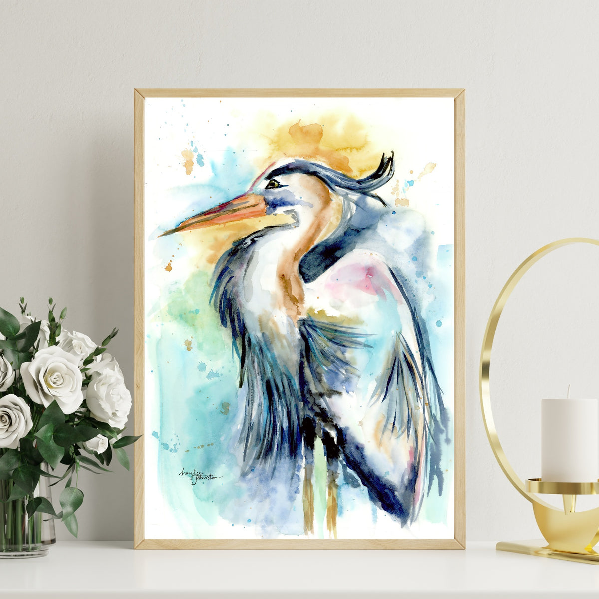 Giclée Female Heron in a slim light wood frame stands on a white tabletop against a pale wall, with a glass vase of white roses to the left and a modern gold ring candle holder with a white pillar candle to the right.