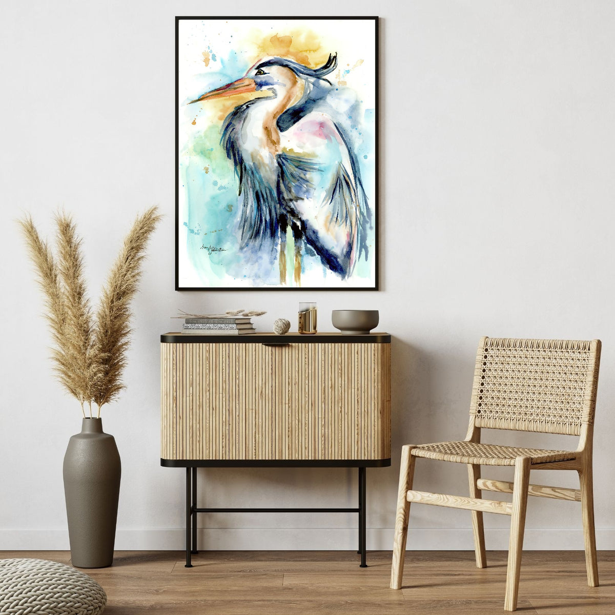 Giclée Female Heron in a slim black frame hangs on a soft white wall above a slatted light wood cabinet with black trim, styled with books and small dishes. A woven cane chair sits to the right, and a tall vase of pampas grass stands to the left on the warm wood floor.