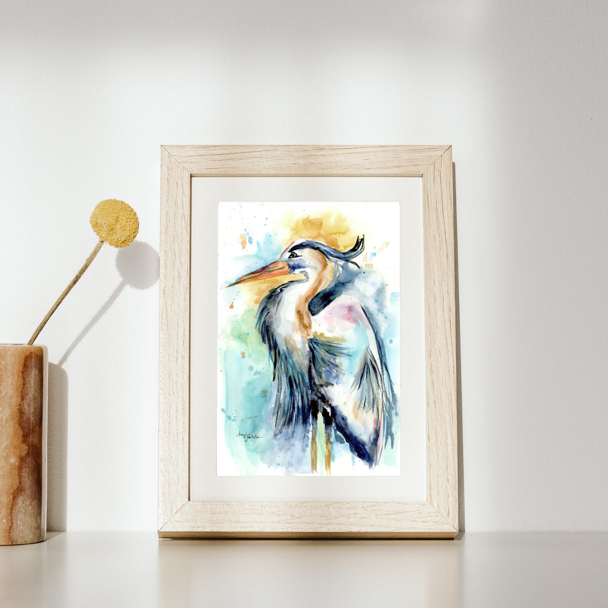 Giclée Female Heron in a natural wood frame with white matting leans against a bright white wall on a white surface, lit by soft sunlight. A stone vase holding a single yellow billy button stem sits to the left.