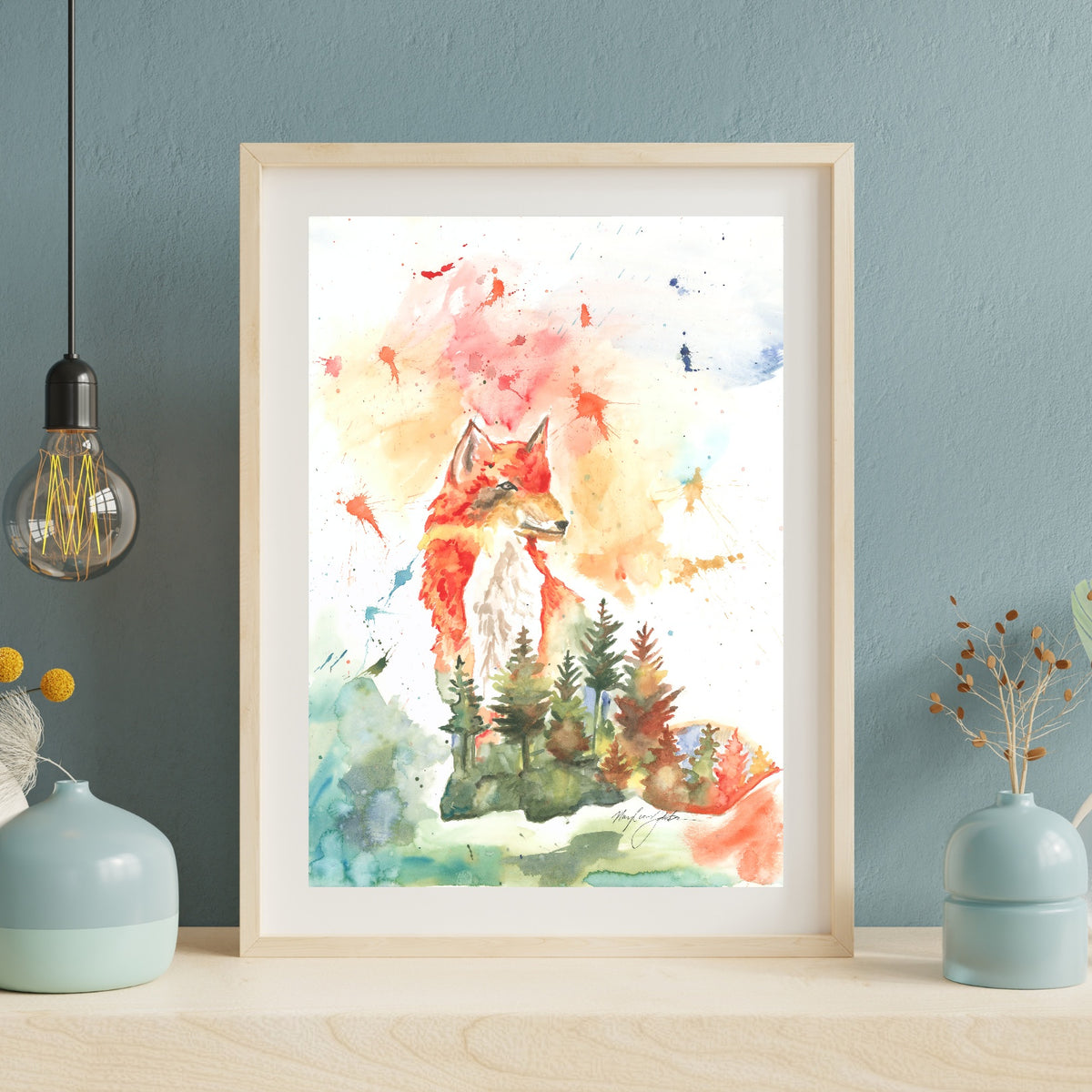 Giclée Forest Fox in a light wood frame with white matting leans against a dusty blue textured wall on a pale wood surface. An exposed Edison bulb pendant hangs to the left, and soft blue ceramic vases with dried botanicals sit on either side.