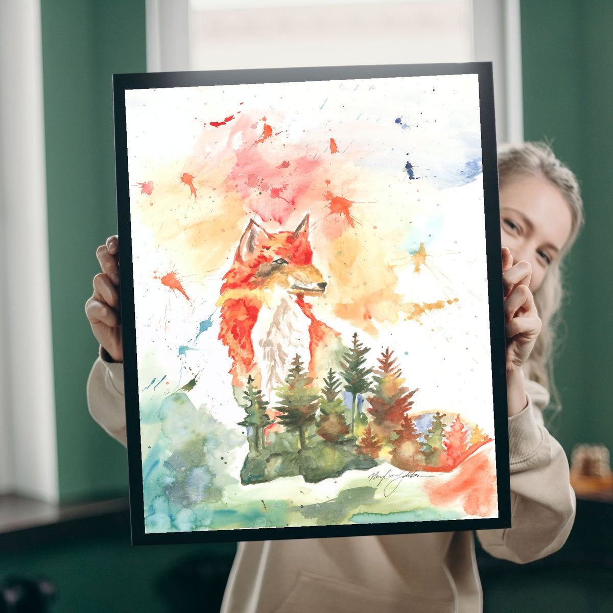 A smiling woman with blonde hair peeks out from behind Giclée Forest Fox, holding it up in a black frame in a room with green walls and bright windows, giving a sense of the print's real-life scale.