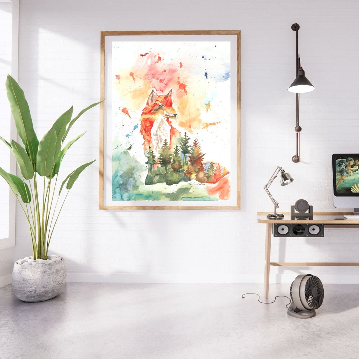 Giclée Forest Fox in a large oak frame with white matting hangs on a white brick wall in a bright studio. A tall leafy plant in a stone planter stands to the left, and a light wood desk with a computer monitor, adjustable lamp, and speaker sits to the right beneath an industrial wall light.
