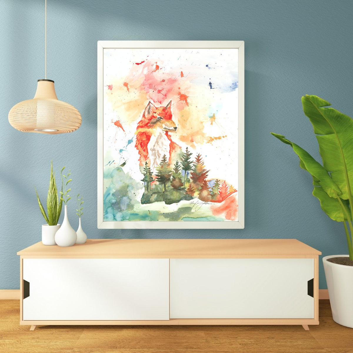 Giclée Forest Fox in a white frame hangs on a soft blue-teal wall above a low light wood console with white sliding doors. A woven rattan pendant lamp hangs to the left, small white vases with greenery sit on the console, and a tall banana leaf plant stands to the right on the warm wood floor.