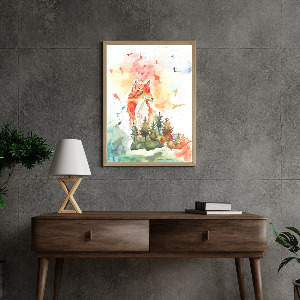 Giclée Forest Fox in a light wood frame hangs on a gray concrete tile wall above a dark walnut mid-century console table. A white lamp with crossed wooden legs and a stack of books sit on the console, with green plants at the edges of the scene.