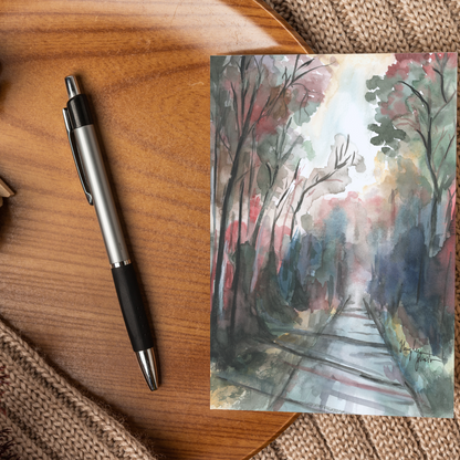 Flat cardstock note card featuring original watercolor piece Forest Road displayed on a wooden surface beside a black and silver pen.
