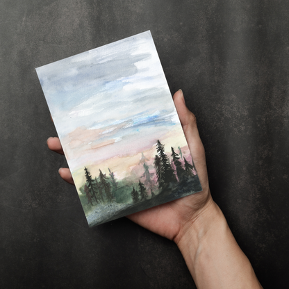 Flat cardstock note card featuring original watercolor piece Forest Skyline in a person's right hand. The background transitions from black at the bottom to a dull gray at the top.