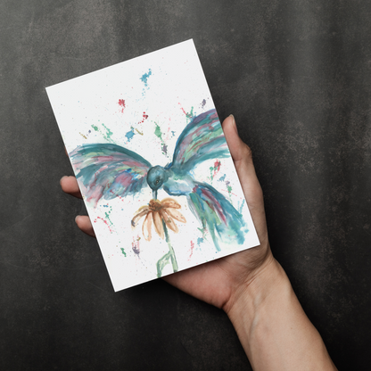 Flat cardstock note card featuring original watercolor piece Hovering Hummingbird in a person's right hand. The background transitions from black at the bottom to a dull gray at the top.