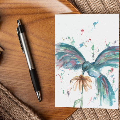 Flat cardstock note card featuring original watercolor piece Hovering Hummingbird displayed on a wooden surface beside a black and silver pen.