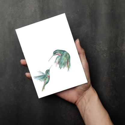 Flat cardstock note card featuring original watercolor piece Hummingbird Duel in a person's right hand. The background transitions from black at the bottom to a dull gray at the top.