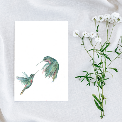Flat cardstock note card featuring original watercolor piece Hummingbird Duel on a white linen background next to fresh white flowers.
