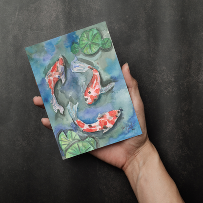 Flat cardstock note card featuring original watercolor piece Koi Pond in a person's right hand. The background transitions from black at the bottom to a dull gray at the top.