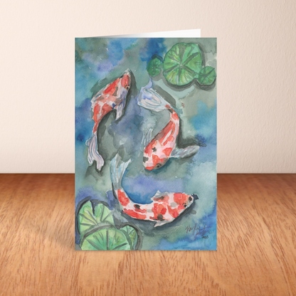 Folding cardstock greeting card featuring original watercolor piece Koi Pond standing upright on a wooden table.
