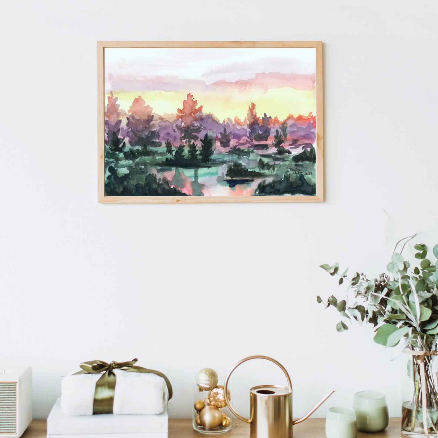 A watercolor painting of a vibrant sunrise over a lake. The sky turns from pink, to yellow, to deep purple in the trees. The water has small clumps of land with a handful of trees reflecting on the water. The watercolor art is in a wooden frame with a table below.