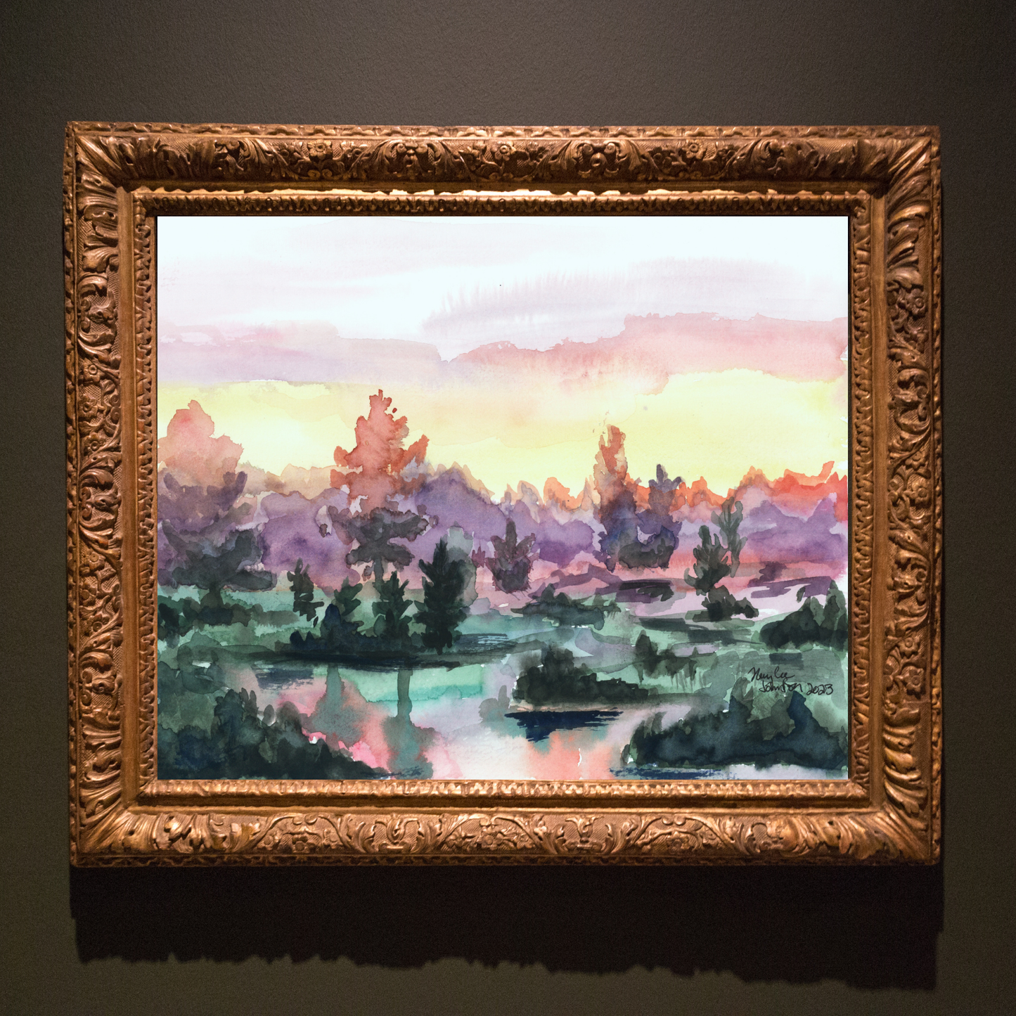 A watercolor painting of a vibrant sunrise over a lake. The sky turns from pink, to yellow, to deep purple in the trees. The water has small clumps of land with a handful of trees reflecting on the water. The painting is in an ornate gold frame on a dark grey background.