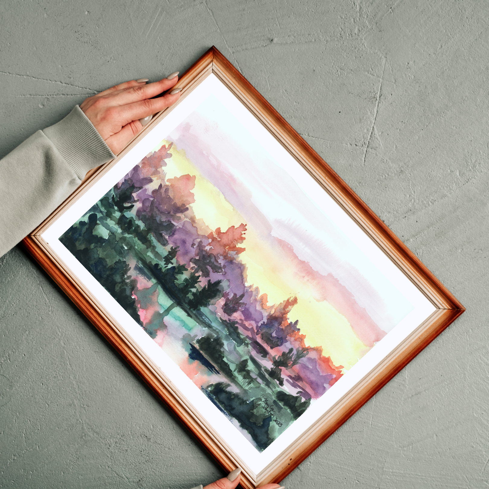 A watercolor painting of a vibrant sunrise over a lake. The sky turns from pink, to yellow, to deep purple in the trees. The water has small clumps of land with a handful of trees reflecting on the water. The piece is framed in a rich red wooden frame against a concrete background. There is a woman whose hands can be seen adjusting the position.