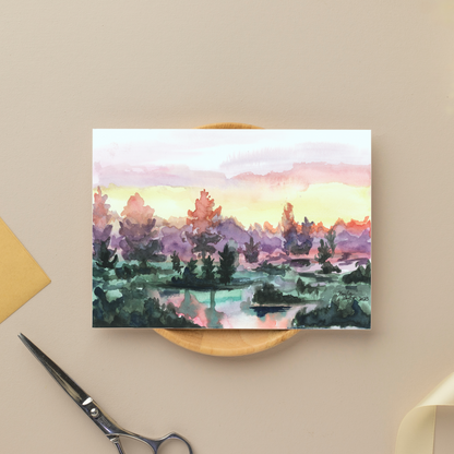Flat cardstock note card featuring original watercolor piece Misty Sunrise Resting on a small wooden plate next to silver scissors.
