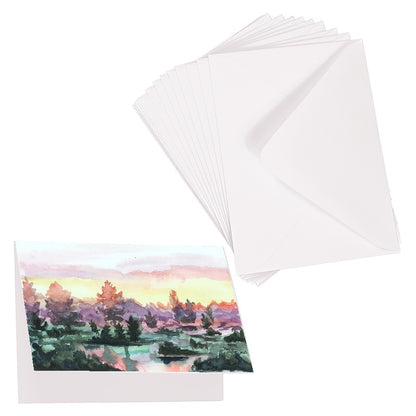 Folding cardstock greeting card featuring original watercolor piece Misty Sunrise laying next to a stack of euro flap envelopes.
