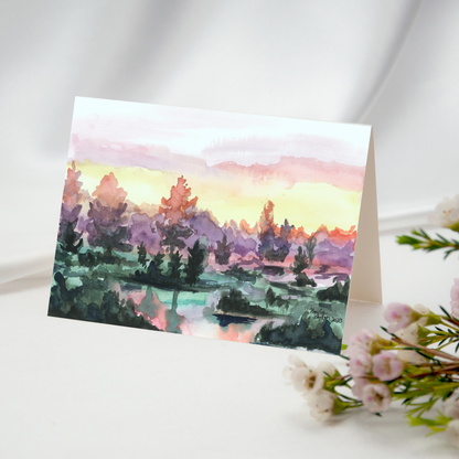 Folding cardstock greeting card featuring original watercolor piece Misty Sunrise resting open on a white tablecloth with pink flowers in the foreground.