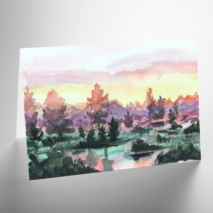Folding cardstock greeting card featuring original watercolor piece Misty Sunrise resting open and backlit on a white table.