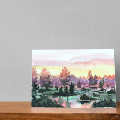 Folding cardstock greeting card featuring original watercolor piece Misty Sunrise resting open on a wooden table against a white wall.