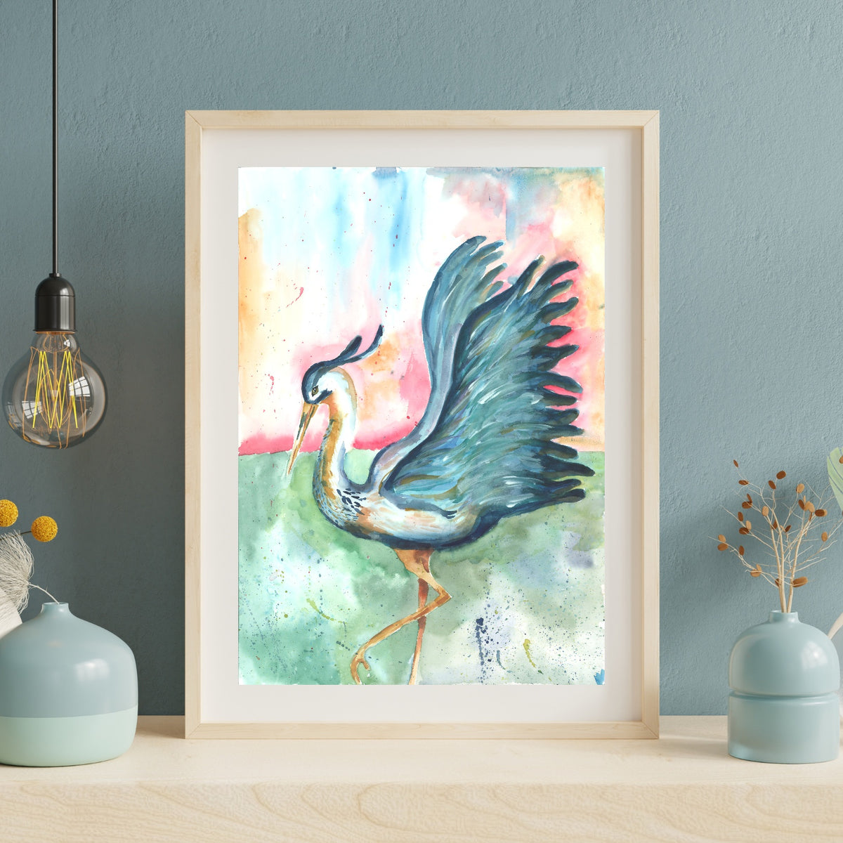 Giclée Morning Glories in a light wood frame with white matting leans against a dusty blue textured wall on a pale wood surface. An exposed Edison bulb pendant hangs to the left, and soft blue ceramic vases with dried botanicals sit on either side.