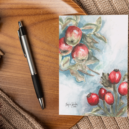 Flat cardstock note card featuring original watercolor piece Red Apples and Rosehips displayed on a wooden surface beside a black and silver pen.