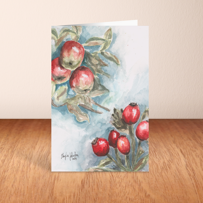 Folding cardstock greeting card featuring original watercolor piece Red Apples and Rosehips standing upright on a wooden table.