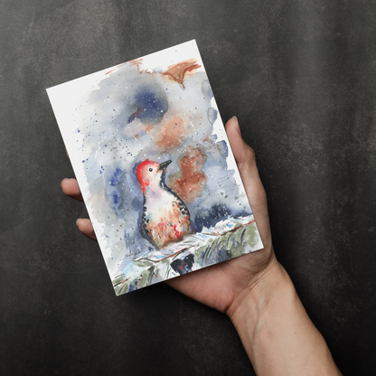 Flat cardstock note card featuring original watercolor piece Red Bellied Woodpecker in a person's right hand. The background transitions from black at the bottom to a dull gray at the top.
