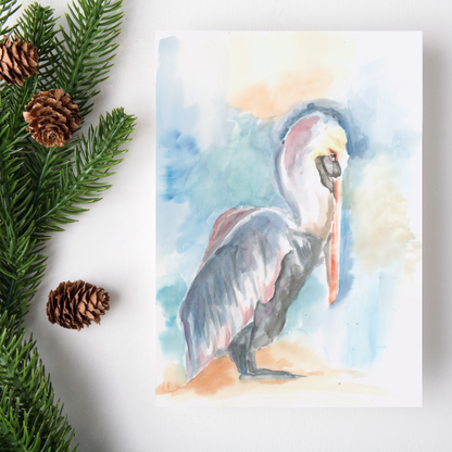 Flat cardstock note card featuring original watercolor piece Shy Pelican on a white background. Green pine fronds with brown pinecones decorate the left side.