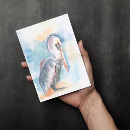 Flat cardstock note card featuring original watercolor piece Shy Pelican in a person's right hand. The background transitions from black at the bottom to a dull gray at the top.
