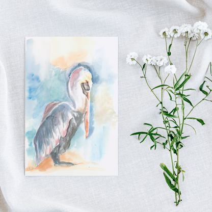 Flat cardstock note card featuring original watercolor piece Shy Pelican on a white linen background next to fresh white flowers.