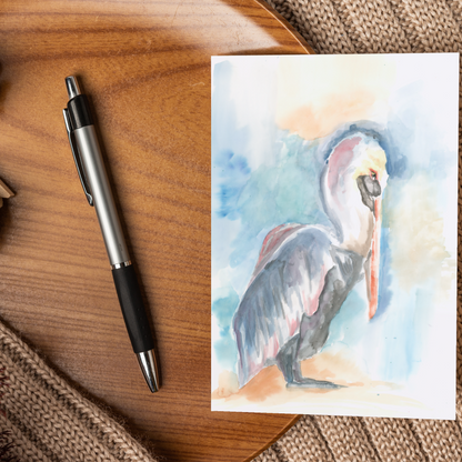 Flat cardstock note card featuring original watercolor piece Shy Pelican displayed on a wooden surface beside a black and silver pen.