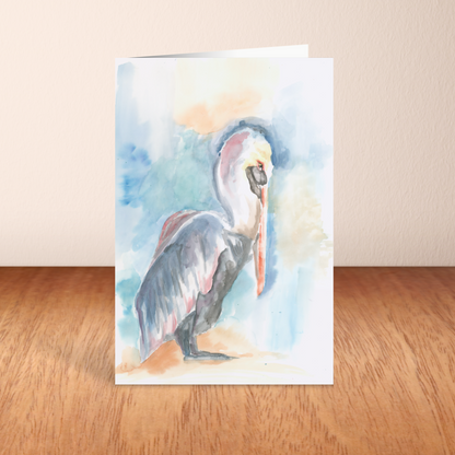 Folding cardstock greeting card featuring original watercolor piece Shy Pelican standing upright on a wooden table.